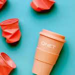 Orange reusable QNET tumbler surrounded by crumpled plastic cups